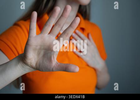 Nahaufnahme eines Porträts einer jungen Frau in einem orangefarbenen Hemd, die ihr Gesicht mit den Händen versteckt, Stress, Angst oder emotionalen Kampf vermittelt. Ideal für psychische Gesundheit, Lebensstil, Wellness und soziale Kampagnen. Stockfoto