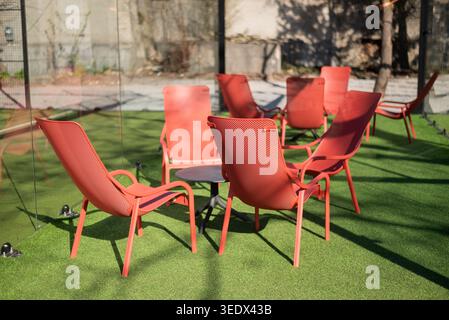 Moderne rote Lounge-Stühle, die um einen Tisch auf künstlichem grünem Gras in einem hellen Terrassenbereich angeordnet sind. Stockfoto