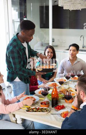 Junger Mann serviert Pasta an Freunden, die an einem Tisch sitzen und Essen und Wein genießen Stockfoto