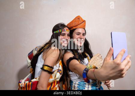 Zwei junge Frauen lächeln und machen ein Selfie in traditioneller Kleidung mit einem Smartphone Stockfoto