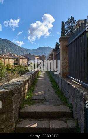 Valldemossa, Spanien - 6. Mai 2026: Ein schmaler Steinweg führt entlang einer alten Steinmauer, auf dem die historische Stadt und die Berge Mallorcas im Hintergrund liegen Stockfoto