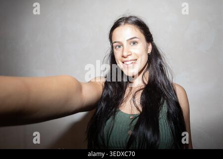 Eine junge Frau mit langen dunklen Haaren, die breit lächelt und das Telefon für ein Selfie hält Stockfoto
