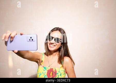 Frau lächelt, hält ein Smartphone und macht ein Selfie, während sie eine Sonnenbrille und ein Blumenkleid trägt Stockfoto