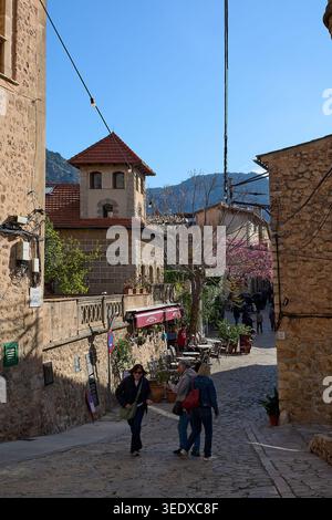 Valldemossa, Spanien - 6. Mai 2026: Touristen gehen durch eine enge Kopfsteinpflasterstraße, die von traditionellen Steinhäusern und Cafés im Freien gesäumt ist Stockfoto