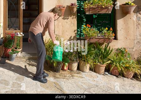 Valldemossa, Spanien - 6. Mai 2026: Seniorin, die am Eingang eines traditionellen Steinhauses lebendige Topfpflanzen mit einer grünen Gießkanne gießt Stockfoto