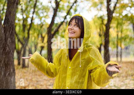 Glückliche junge Frau in gelbem Regenmantel genießt Herbstregen im grünen Park draußen Stockfoto