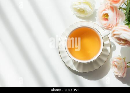 Elegante Lifestyle-Komposition mit weißer Keramik-Teetasse mit Muscheluntertasse und goldenem Rand, gefüllt mit bernsteinfarbenem grünem Tee, umgeben von blassrosa Stockfoto