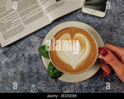 Warmer, cremiger Latte in den Händen gehalten mit eleganter Herzlattekunst auf dunklem, strukturiertem Hintergrund. Stockfoto