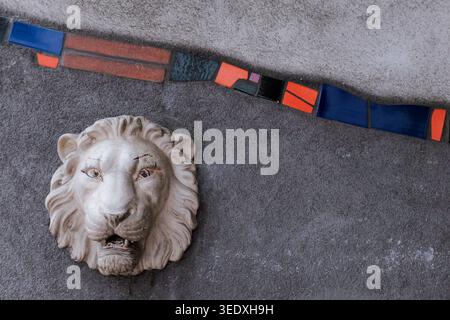 Wien, Österreich: Nahaufnahme einer weißen Stein-Löwenkopf-Skulptur auf grauer texturierter Wand mit farbenfroher Mosaikfliese. Architekturhintergrund. Stockfoto