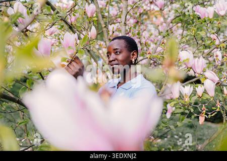 Selbstbewusste Afrikanische Frau, Die Zwischen Rosa Magnolienbäumen Steht Und Nachdenklich Wegblickt Stockfoto