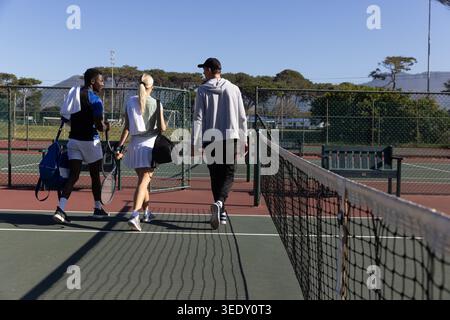 Verschiedene Freunde laufen auf dem Tennisplatz in Richtung Tor mit Schlägern, Taschen und Handtüchern in Sportkleidung. Outdoor, Sport, Gruppe, Kameradschaft, Sonnenschein, Stockfoto