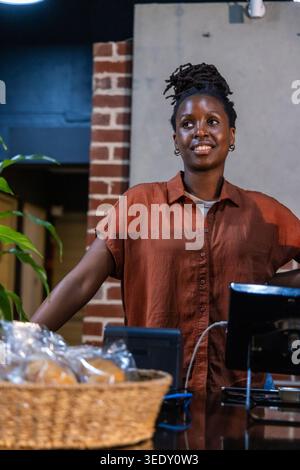 Afrikanische Frau lächelt und steht hinter der Theke in der Bäckerei, trägt Rosthemd, gewebten Korb, POS. Cafe, Gastfreundschaft, gemütlich, Ziegelstein, Pflanze, Personal, Pastor Stockfoto