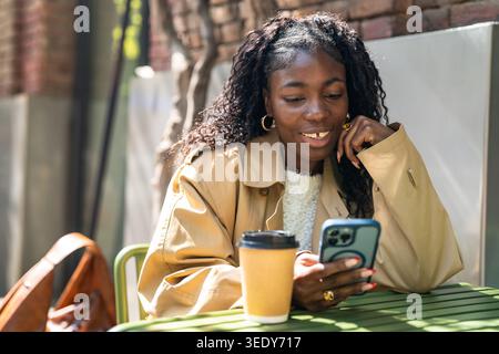 Fröhliche afroamerikanische Frau, die auf dem Smartphone scrollt, während sie sich am warmen Frühlingstag im Café entspannt. Stockfoto