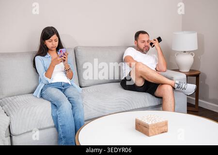 Teenager und Vater ignorieren sich gegenseitig, während sie auf ihren Mobiltelefonen surfen. Eine Szene digitaler Ablenkung und fehlender Kommunikation in den Lebenden Stockfoto