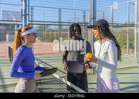 Eine Gruppe von Freunden, die auf einem Pickleball-Platz geselligt und lacht, während sie Paddel und Ball halten, während sie den Sport diskutieren und Outdoor-Aktivitäten genießen Stockfoto
