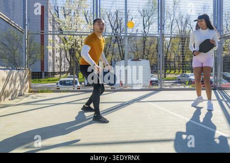Zwei junge, vielfältige Freunde spielen energiegeladene Pickleball auf einem sonnigen städtischen Platz, lächeln und aktiv, während sie sich mit Paddeln zusammentun und Fitness, Spaß und Spaß genießen Stockfoto