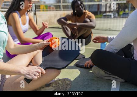 Vielfältige Freunde, die sich auf einem Freiluftplatz entspannen, Pickleball-Ausrüstung halten und nach einem aktiven Spiel gesellige Zeit genießen, symbolisieren Gemeinschaft, Fitne Stockfoto