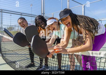 Verschiedene Freunde lehnen sich spielerisch über ein Pickleball-Netz, lachen und halten Paddel und genießen gemeinsam ein aufregendes und unterhaltsames Outdoor-Sportspiel auf einem Stockfoto