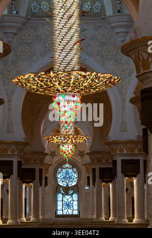Blick auf einen riesigen, reich verzierten Kronleuchter mit bunten Kristallen, die von einer gewölbten Decke hängen, in einer großen Moschee mit Marmorsäulen und Blumenmuster Stockfoto