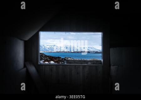 Blick auf das Innere eines verlassenen Bauernhauses mit Blick durch ein Fenster auf schneebedeckte Berge und das blaue Meer in Hafnarfjordur, Hafnarfjardark Stockfoto