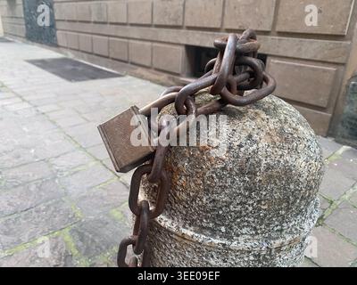 Eine rostfreie Metallkette und ein Vorhängeschloss aus Messing sichern einen verwitterten Granitpfoller, die Konzepte der Sicherheit, der Einschränkung und der städtischen Infrastruktur in darstellen Stockfoto