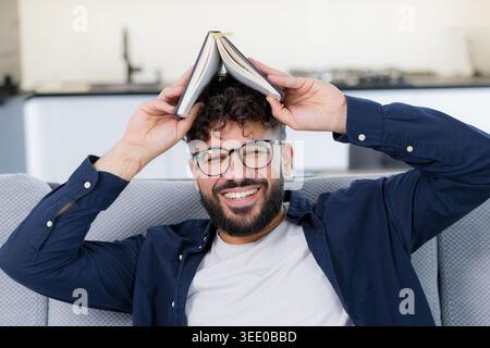 Junger Mann mit einer Brille, der breit lächelt, ein offenes Notizbuch wie ein Dach auf seinen lockigen Haaren hält und sich auf einem Sofa entspannt Stockfoto