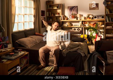 Junger erwachsener kaukasischer Mann, der auf dem Bett sitzt, sich nach dem Aufwachen im Schlafzimmer dehnt und den Tag beginnt. Nützlich für Schlaf Gesundheit, Morgen Lifestyle, Work Life Balance Themen Stockfoto