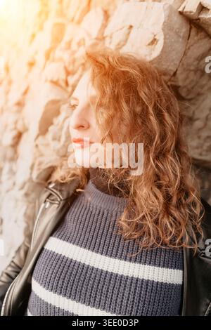 Frau lehnende Wand schöne junge Frau mit lockigem Ingwerhaar, die eine Lederjacke und einen gestreiften Pullover gegen eine rustikale Wand trägt Stockfoto
