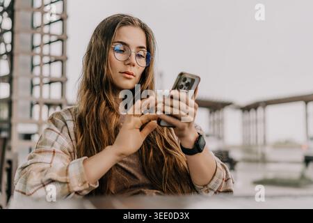 Smartphone-Frau im Freien mit einem mobilen Gerät, während sie im Café im Freien sitzt und eine stilvolle Brille und ein kariertes Hemd trägt Stockfoto