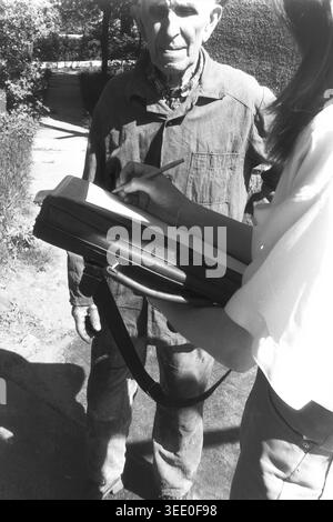Sloviansk, 1997, Stadtzentrum. Ein archiviertes Schwarzweißfoto, das ein Interview im Freien in sonniger urbaner Umgebung zeigt. Ein älterer Mann mit flacher Mütze steht geduldig, während eine junge Frau mit einer großen schwarzen Mappe schriftliche Notizen auf einem Blatt Papier macht. Die Interaktion findet auf einem gepflasterten Gehweg statt, der von Büschen und Bäumen umgeben ist, und fängt einen Moment aktiven Journalismus, Sozialforschung oder Gemeindevermessung ein, der typisch bürgerliches Engagement und das tägliche Leben im friedlichen Donbass reflektiert. Stockfoto