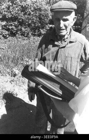 Sloviansk, 1997, Stadtzentrum. Ein archiviertes Schwarzweißfoto, das ein Interview im Freien in sonniger urbaner Umgebung zeigt. Ein älterer Mann mit flacher Mütze steht geduldig, während eine junge Frau mit einer großen schwarzen Mappe schriftliche Notizen auf einem Blatt Papier macht. Die Interaktion findet auf einem gepflasterten Gehweg statt, der von Büschen und Bäumen umgeben ist, und fängt einen Moment aktiven Journalismus, Sozialforschung oder Gemeindevermessung ein, der typisch bürgerliches Engagement und das tägliche Leben im friedlichen Donbass reflektiert. Stockfoto