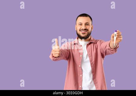 Schöner junger Mann mit Thermometer, das die Daumen-nach-oben-Geste auf lilafarbenem Hintergrund zeigt Stockfoto