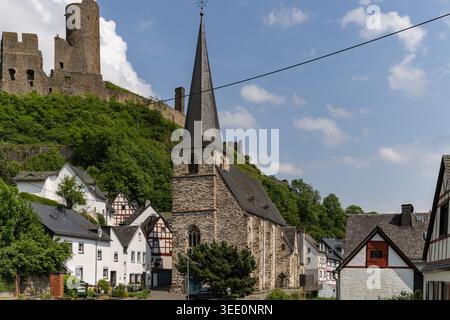 Blick auf die Dreifaltigkeitskirche und Fachwerkhäuser mit mittelalterlichen Burgruinen auf einem üppig grünen Hügel in Monreal, Rheinland-Pfalz, Deutschland. Stockfoto