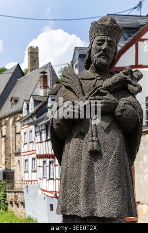 Blick auf die Steinstatue des heiligen Johannes von Nepomuk mit traditionellen Fachwerkhäusern und einem mittelalterlichen Burgturm unter einem blauen Himmel in Monreal, Rhinela Stockfoto