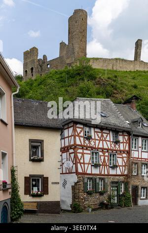 Blick auf traditionelle Fachwerkhäuser im historischen Dorfkern mit den mittelalterlichen Burgruinen auf einem üppig grünen Hügel unter bewölktem Himmel in Monr Stockfoto