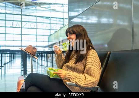 Junge Frau genießt gesunden Salat, während sie am Flughafenterminal wartet Stockfoto