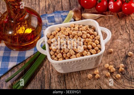 Nahaufnahme Stillleben, getrocknete Kichererbsen in weißer Keramikschale auf rustikalem Holztisch. Zusammensetzung enthält eine Flasche Olivenöl, frische rote Tomaten, Knoblauch, Stockfoto