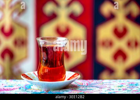 Birnenförmiges Armudu-Glas mit schwarzem Tee auf einer dekorierten Untertasse, mit einem traditionellen aserbaidschanischen Teppich im Hintergrund, Aserbaidschan. Stockfoto