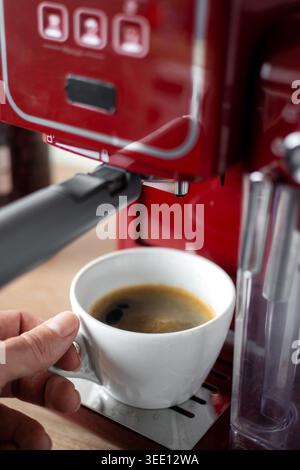 Nahaufnahme einer Person mit einer weißen Tasse gefüllt mit frischem Espresso aus einer roten Espressomaschine in einer modernen Küche. Gemütliche Kaffeezubereitungen zu Hause Stockfoto