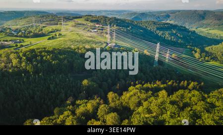 Luftaufnahme von Hochspannungsleitungen durch grüne Wälder und Hügel in Deutschland, Energieübertragungsinfrastruktur in ländlicher Landschaft bei Sonnenuntergang Stockfoto
