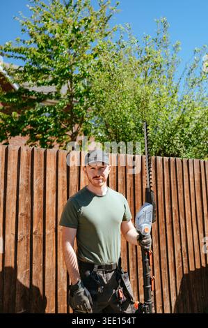 Mann, der eine Heckenschere hält und am Holzzaun im Garten steht Stockfoto