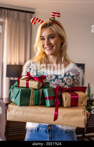 Frau in ihren 20ern, die zu Hause eingewickelte Geschenke hält, einen Fuzzy-Pullover und ein Zuckerrohr-Stirnband trägt. Weihnachten, Geschenke, festlich, gemütlich, warm, Beleuchtung, Wohnzimmer Stockfoto