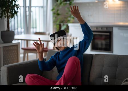 Weiße Frau, die VR-Brillen für Unterhaltung und Spiele mit Augmented-Reality-Technologie verwendet. Mädchen sitzt auf dem Sofa mit futuristischem Headset auf den Augen, um Videos mit AR-Funktion anzusehen Stockfoto