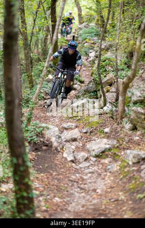 Ein Mann fährt aggressiv auf einem schmalen, felsigen Waldweg mit Schutzausrüstung und Action-Kamera, während andere Fahrer hinter ihm zwischen Bäumen und Felsen verschwimmen. Stockfoto