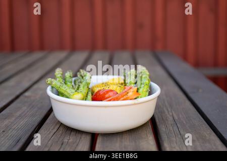Gegrilltes Spargelmais und Tomatengemüse, serviert in einer Schüssel auf rustikalem Holztisch im Freien Stockfoto