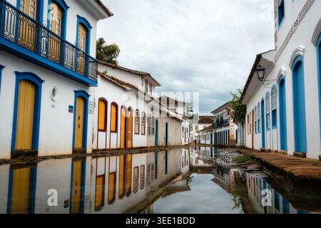 Blick auf historische Kolonialgebäude mit bunten Türen und Fenstern, die sich in einer überfluteten Kopfsteinpflasterstraße unter einem bewölkten Himmel in Paraty, Bundesstaat R, spiegeln Stockfoto