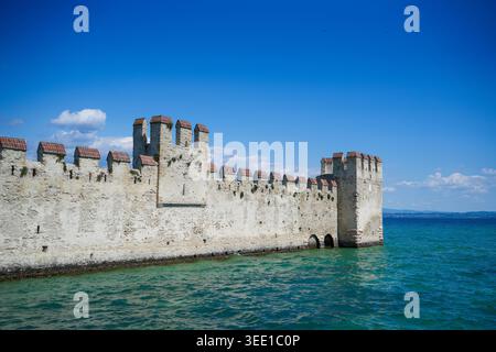 Blick auf die historischen Mauern und Türme der Burg Scaligero, die sich aus dem türkisfarbenen Wasser des Gardasees unter einem klaren blauen Himmel in Sirmione, Lombardei, erheben Stockfoto