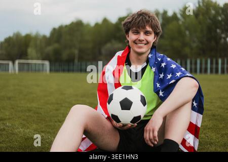 Lächelnder Fußballspieler, der in eine amerikanische Flagge gehüllt ist und einen Fußball auf einem Feld hält. Vorstellung von Sportsieg, Patriotismus, Nationalmannschaft. Stockfoto