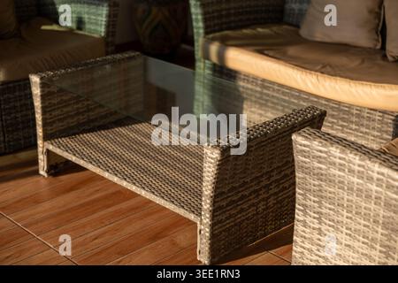 Couchtisch aus Korb mit Glasplatte und Rattan-Stuhl auf der Terrasse schaffen einen entspannenden, komfortablen Sitzbereich im Freien, der Sonnenlicht auf warmem, friedlichem A reflektiert Stockfoto