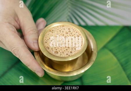 Hand mit frischem, heißem Milchtee Chai in einer Metalltasse. Traditioneller indischer Masala Chai-Tee zum Frühstück in einem Messing-Kaffeeglasbecher, Blick von oben. Niemand Stockfoto